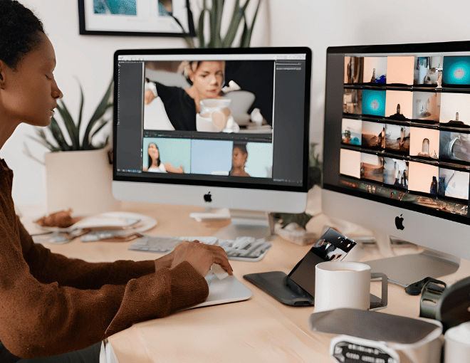 A video editor meticulously adjusts clips on a Mac desktop, surrounded by dual monitors and coffee cups.