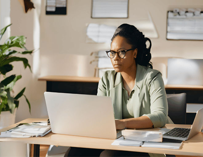 A woman with eyeglasses types on her laptop, focused intently, seated at a desk with a coffee mug beside her.
